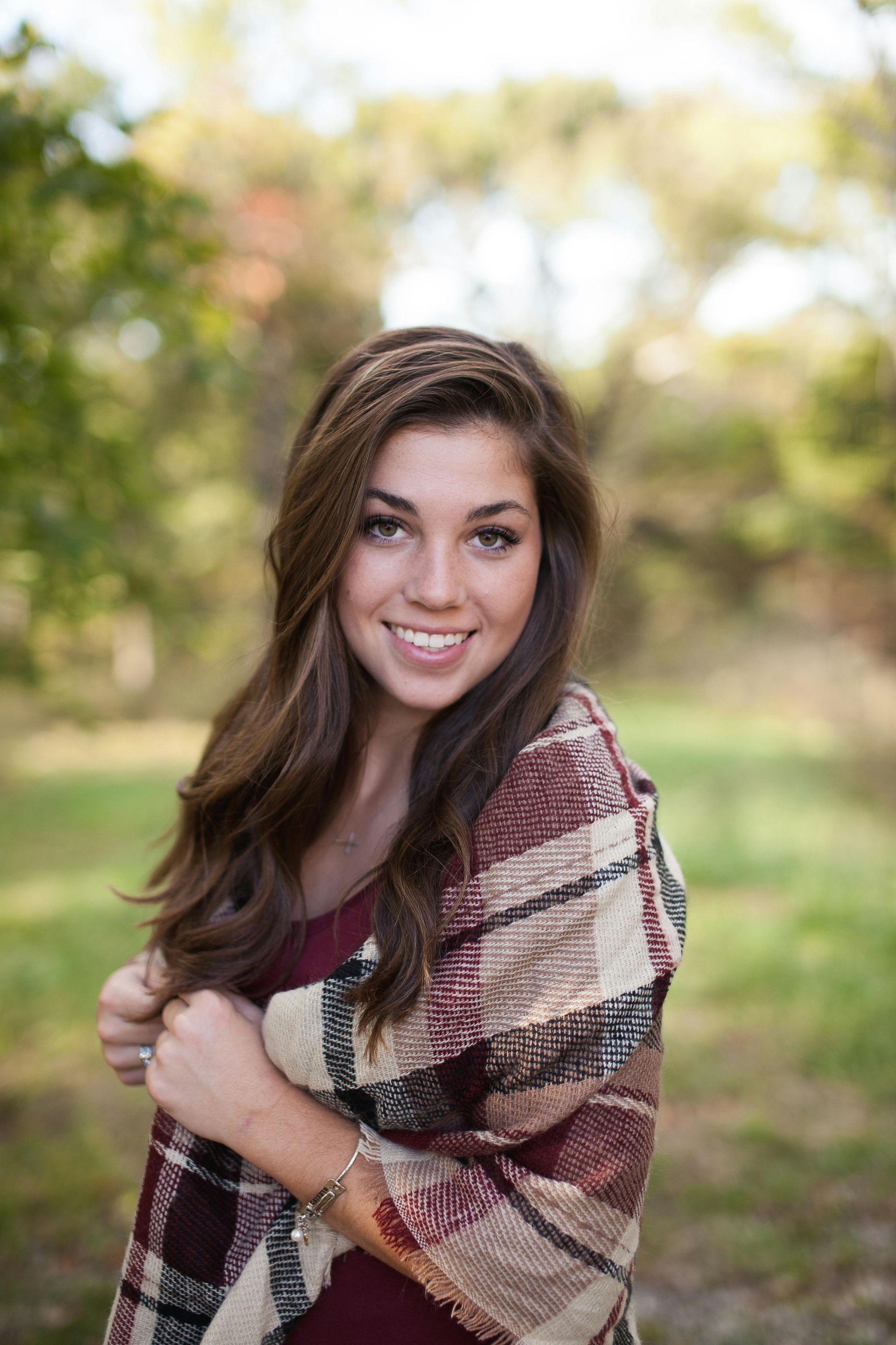 Woman smiling, wrapped in a plaid shawl, standing outdoors in front of blurred trees.