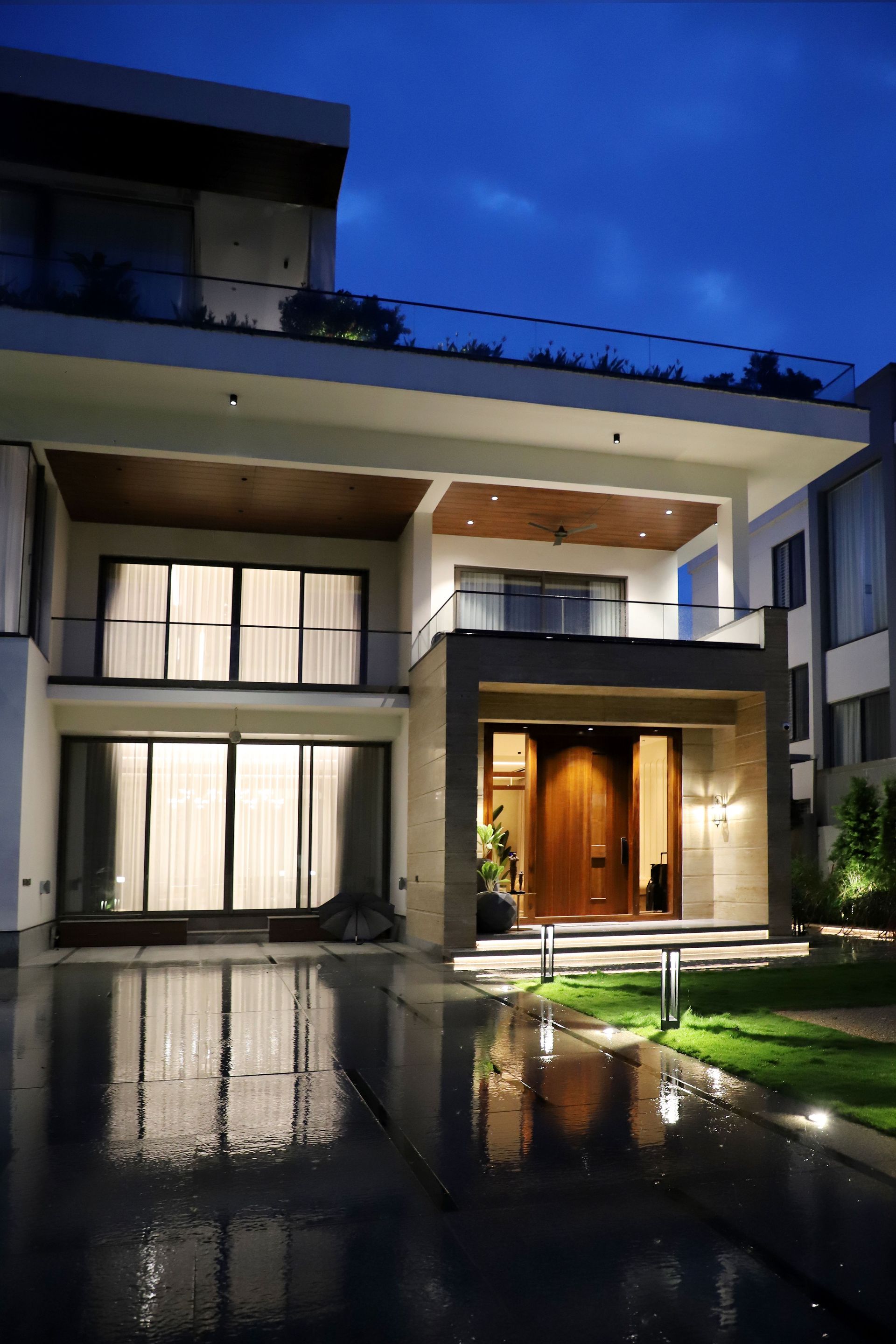 Modern two-story house at dusk with warm lights and reflections on a wet driveway