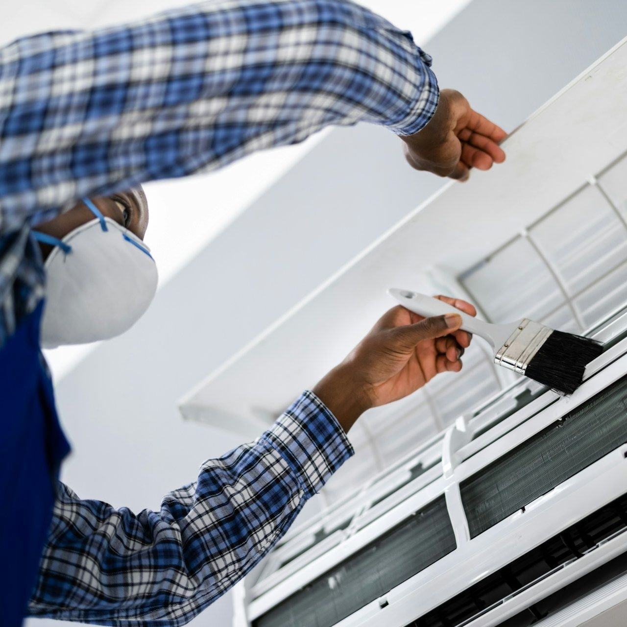 Technician cleaning white wall-mounted air conditioner with a brush while wearing a blue plaid shirt and mask
