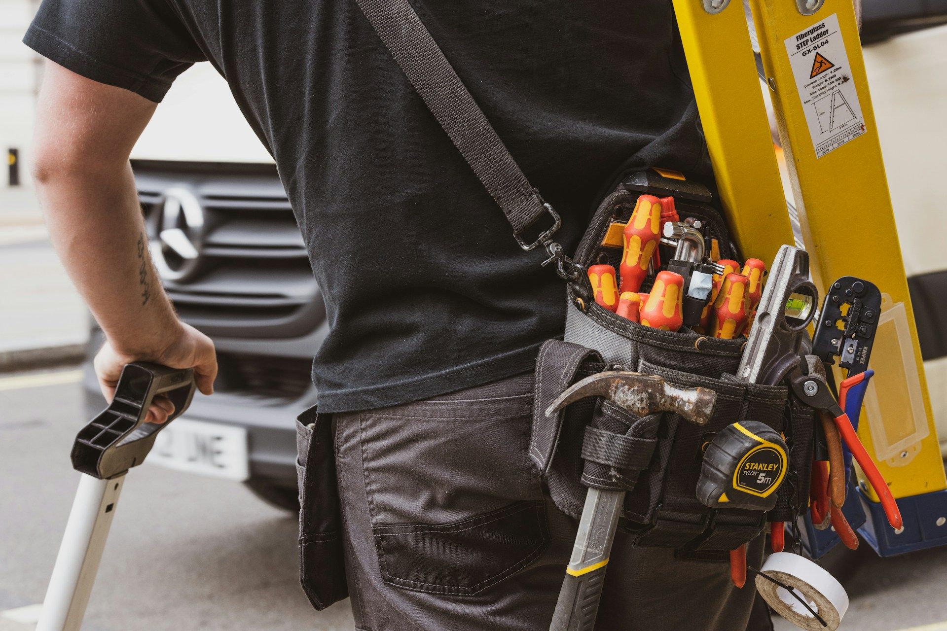 Worker in black shirt with tool belt beside a yellow utility truck, holding a pry bar.
