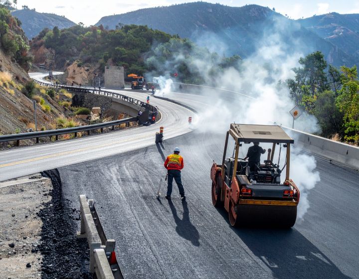 Road paving in progress on a winding mountain road; steam rises; workers and heavy machinery are present.