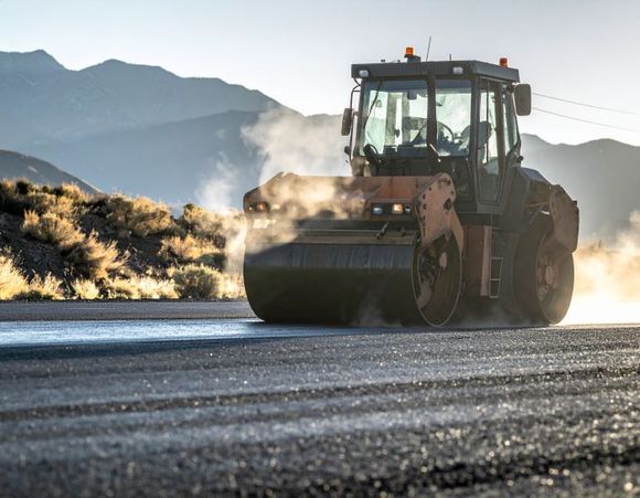 Road roller compacting fresh asphalt on a road, mountains in the background.