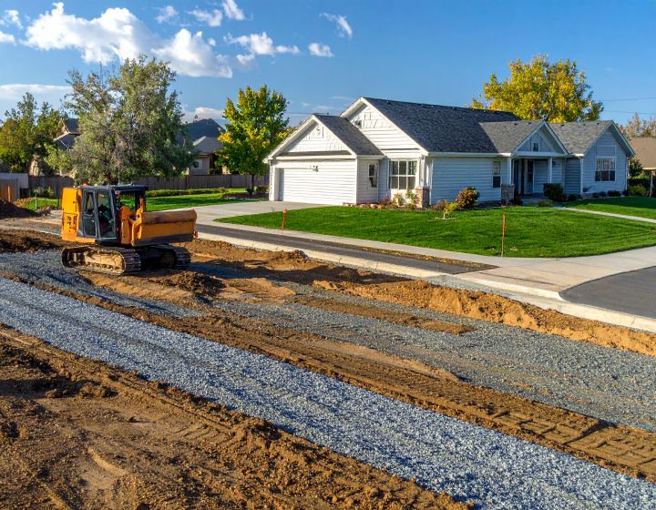 Yellow excavator laying gravel along a curb, next to a white house with a green lawn.