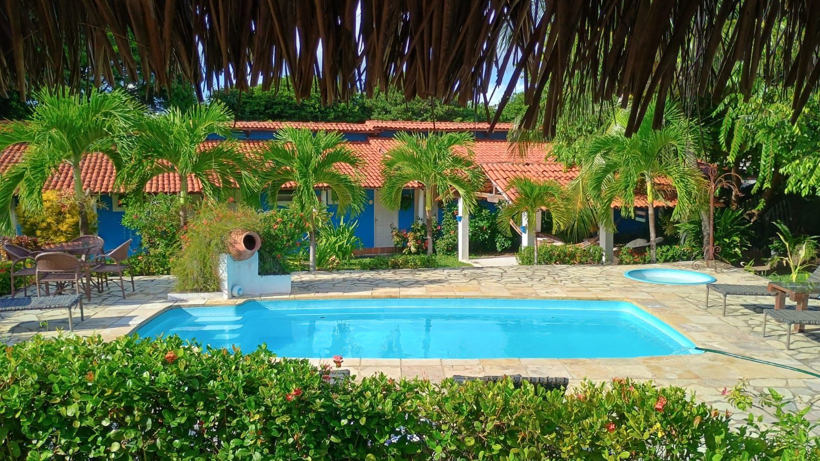 Piscina em frente a um prédio azul com telhado laranja, rodeada por palmeiras e vegetação exuberante.