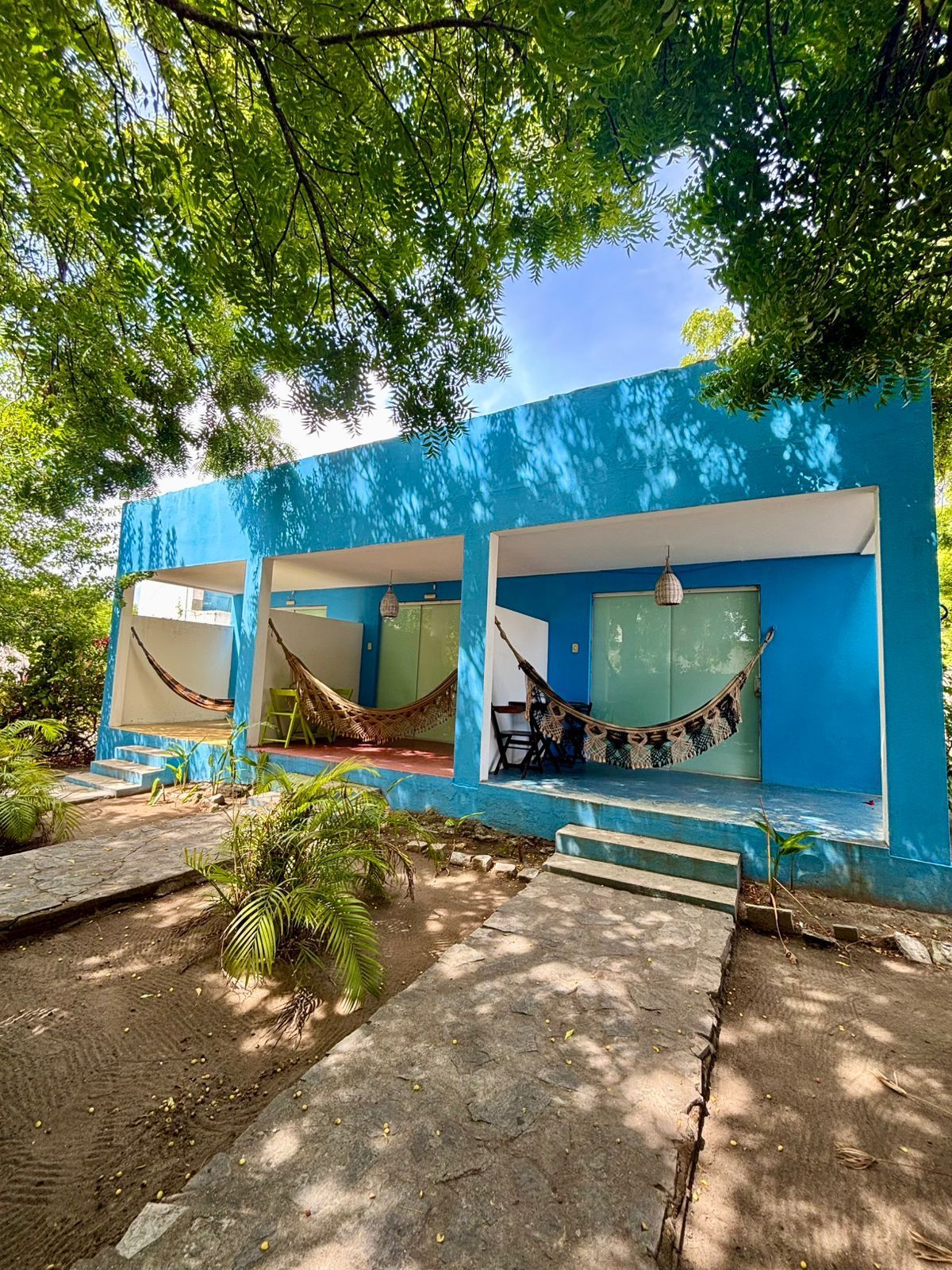 Prédio azul com redes em um dia ensolarado. Caminho de concreto leva aos quartos ao ar livre.