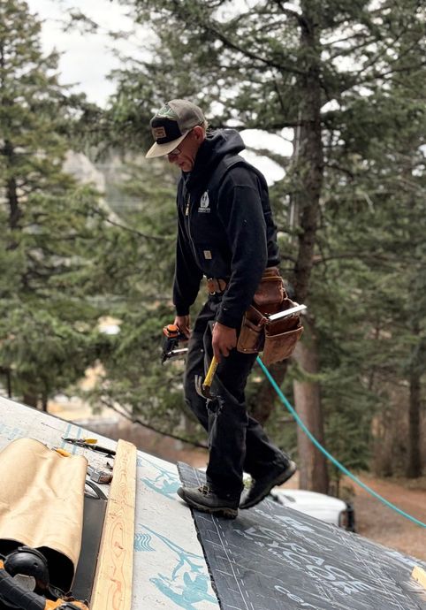 A worker wearing a cap and tool belt walks across a sloped roof underlayment with trees in the background.