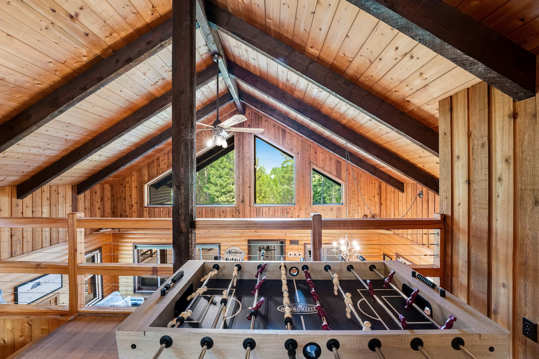 A foosball table sits in a rustic wooden loft with a vaulted ceiling and large windows overlooking the room below.