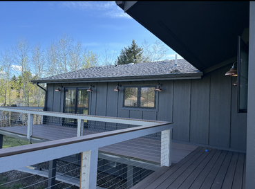 A modern house exterior with dark vertical siding, a spacious wooden deck with a white railing, and a gray shingled roof.