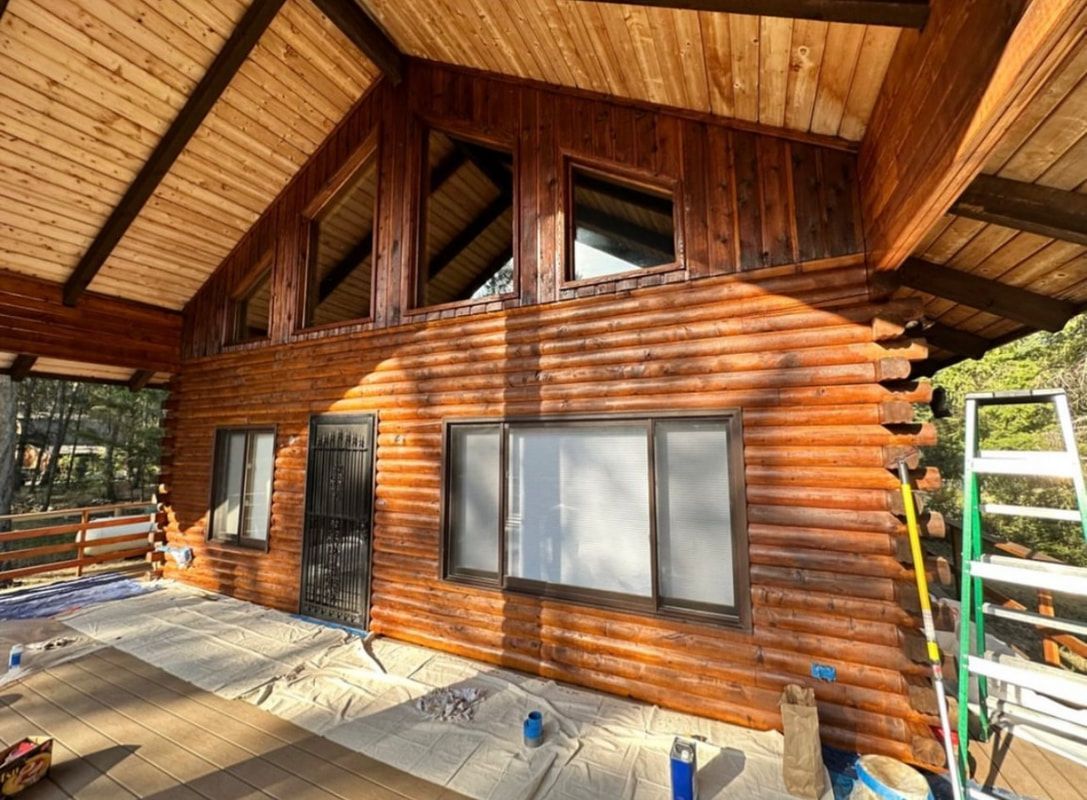A rustic log cabin exterior with a large window, dark wood trim, and an A-frame roof during a home renovation.