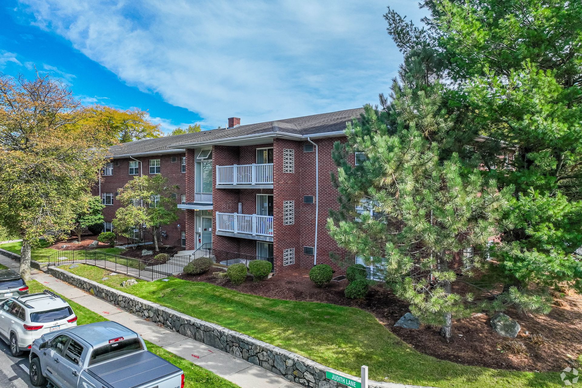 Brick apartment building with balconies beside trees and parked cars on a sunny day at Waltham Overlook Apartments in Waltham, MA.