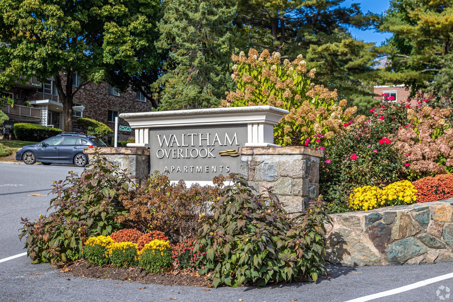 Stone entrance sign for Waltham Hall with landscaped flowers and trees beside a parking lot at Waltham Overlook Apartments in Waltham, MA.