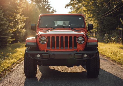 Red Jeep Wrangler parked on a sunlit country road surrounded by trees and warm afternoon light.
