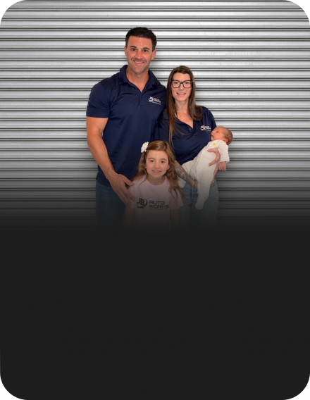 Three people posing in front of a gray corrugated metal wall, one holding a baby | JB Auto Works