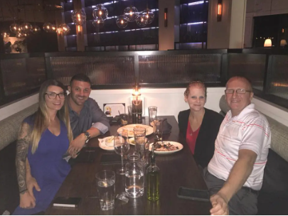 Four people seated at a restaurant table, smiling with drinks and plates in a dimly lit setting.