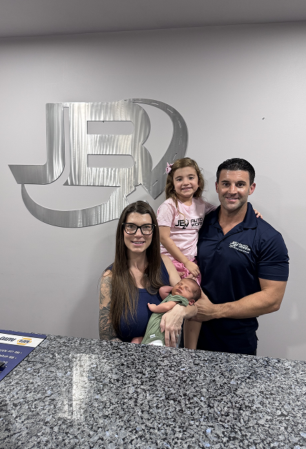 Family of three smiling at a reception desk with a silver logo on the wall behind them. | JB Auto Works