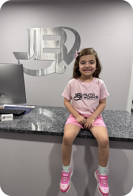 Girl in pink outfit sitting on a counter, smiling in front of a JEB sign. | JB Auto Works