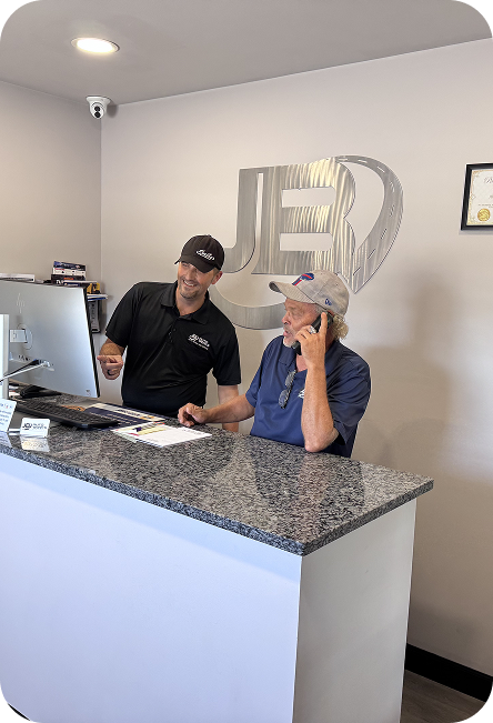 Two people talking at a reception desk in a bright office lobby, with a white wall logo behind them. | JB Auto Works