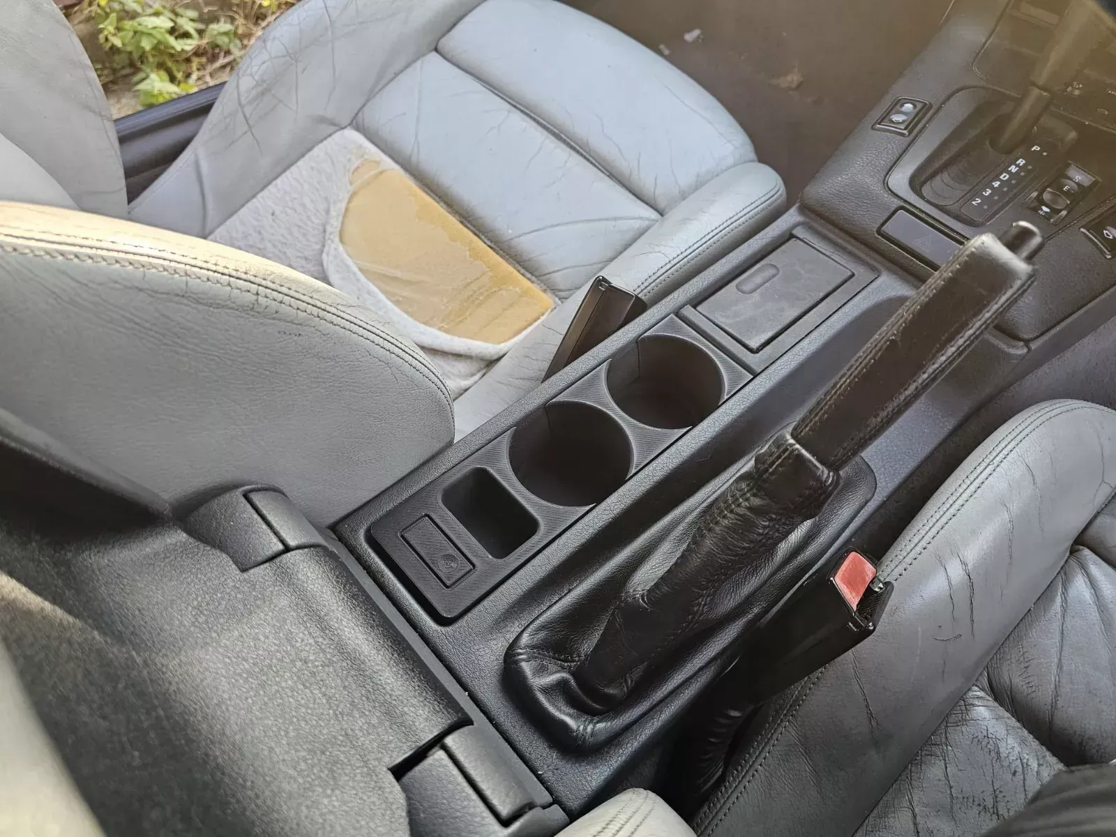 Interior car detail: center console with cup holders, gray leather seats, and a black handbrake.