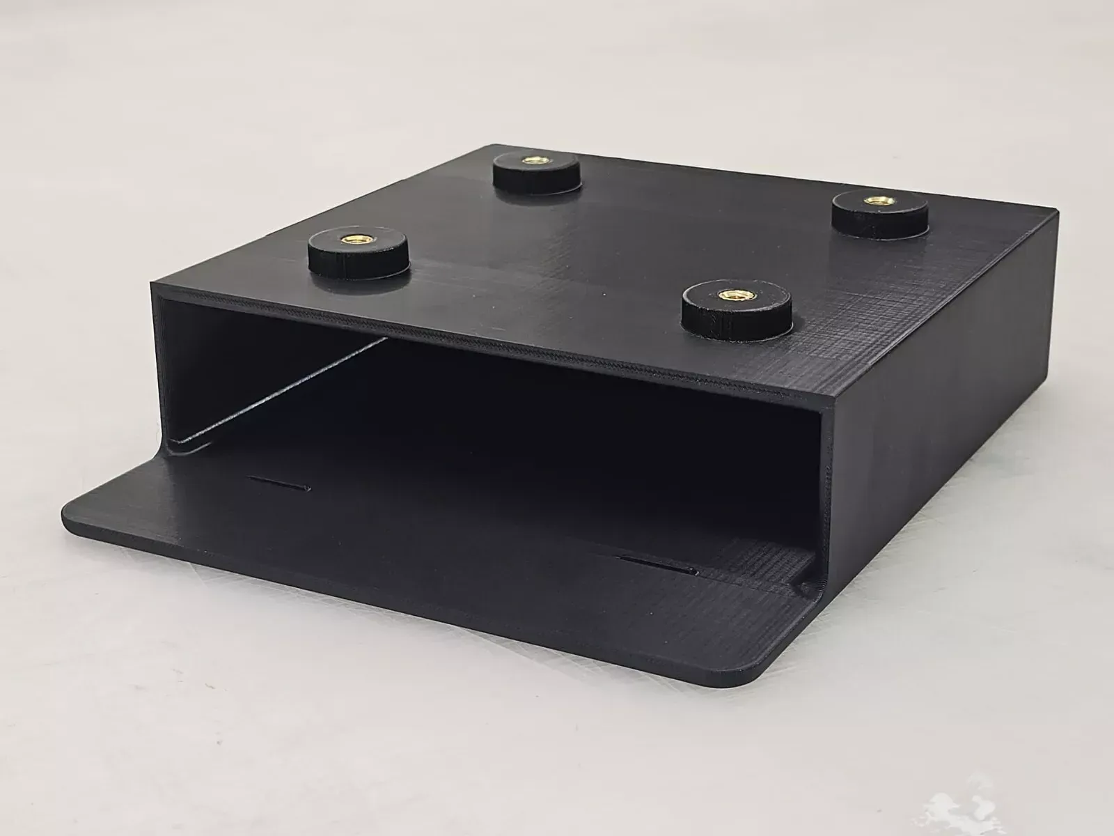 Black 3D-printed box with four brass-colored screws, likely for mounting, on a white surface. A 3D printer is in the blurred background.