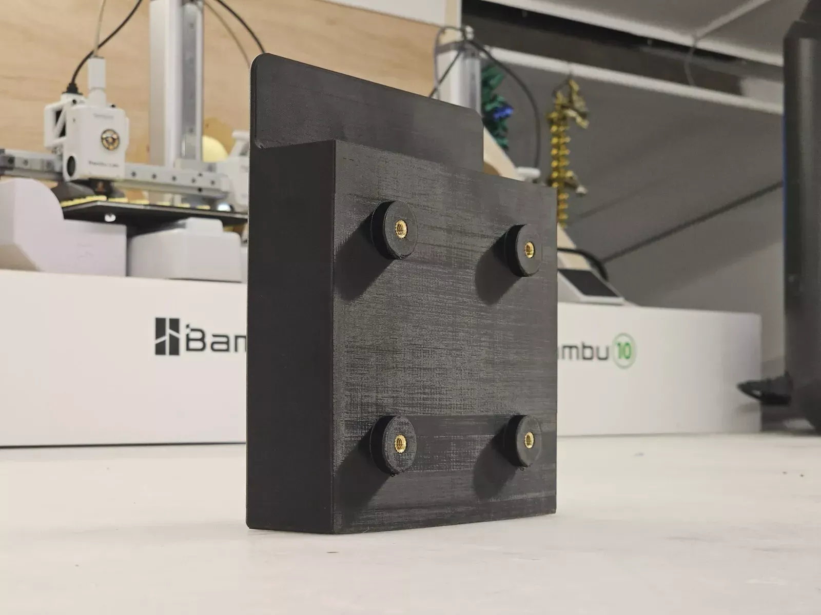 Black 3D-printed box with four brass-colored screws, likely for mounting, on a white surface. A 3D printer is in the blurred background.