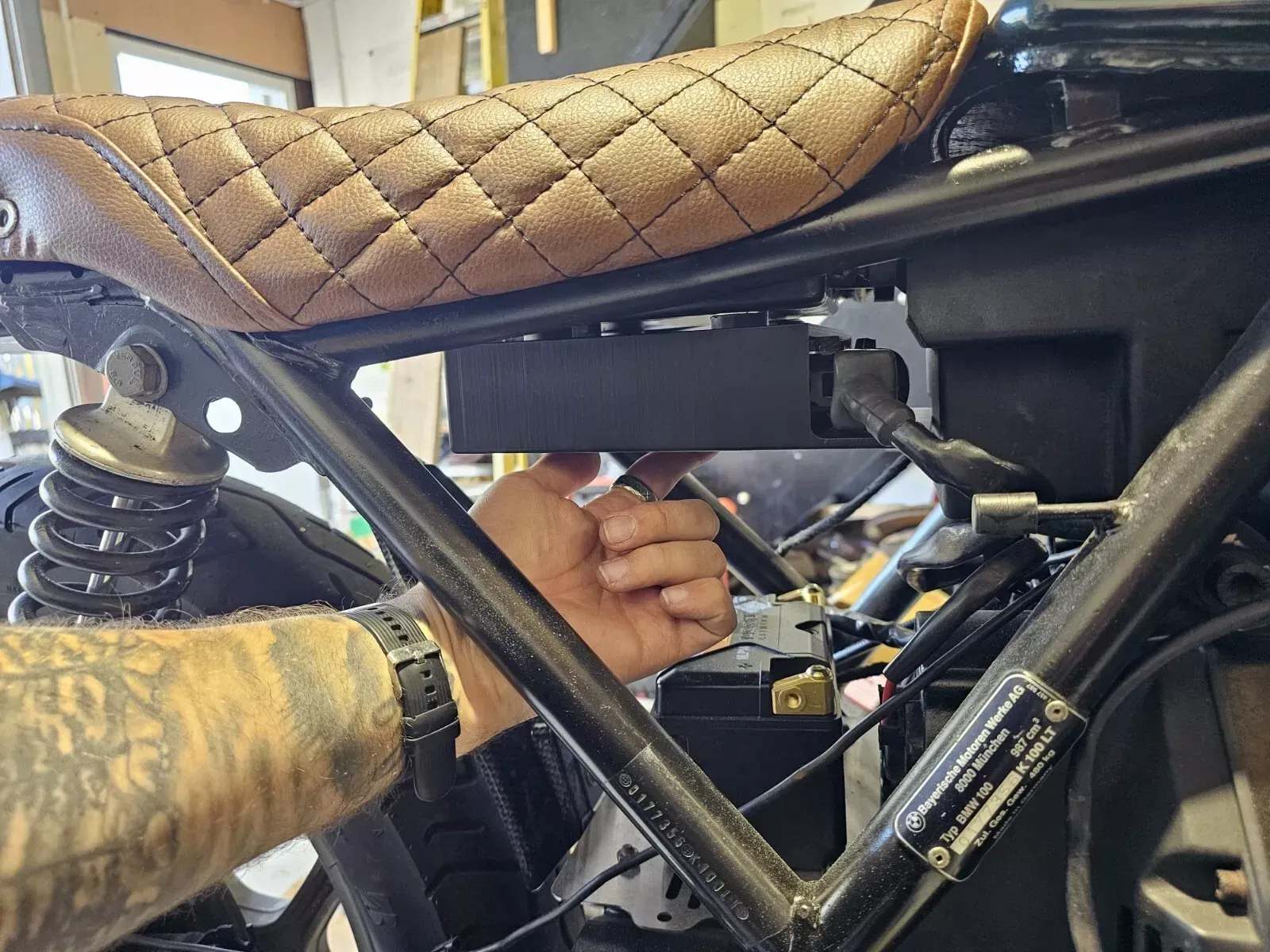 Person working on motorcycle, adjusting black box under brown seat, in a garage.