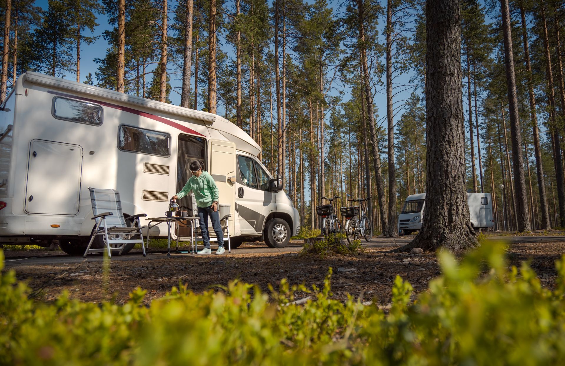 RV parked in a forest, a person stands outside, setting up chairs. Sunlight filters through trees.