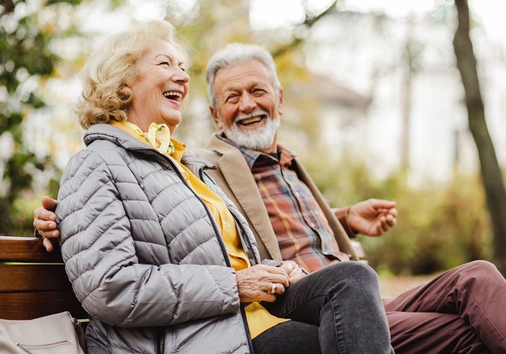 Two smiling people sit on a park bench, laughing together in gray and brown jackets.