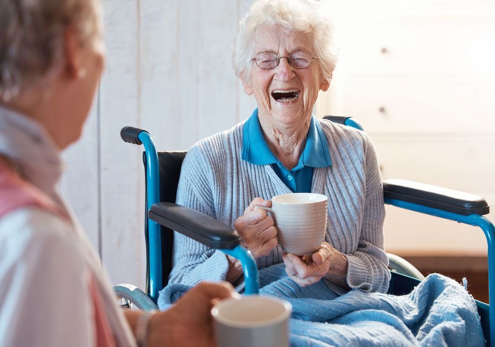 Smiling person in a wheelchair holding a mug and talking with someone in a bright room
