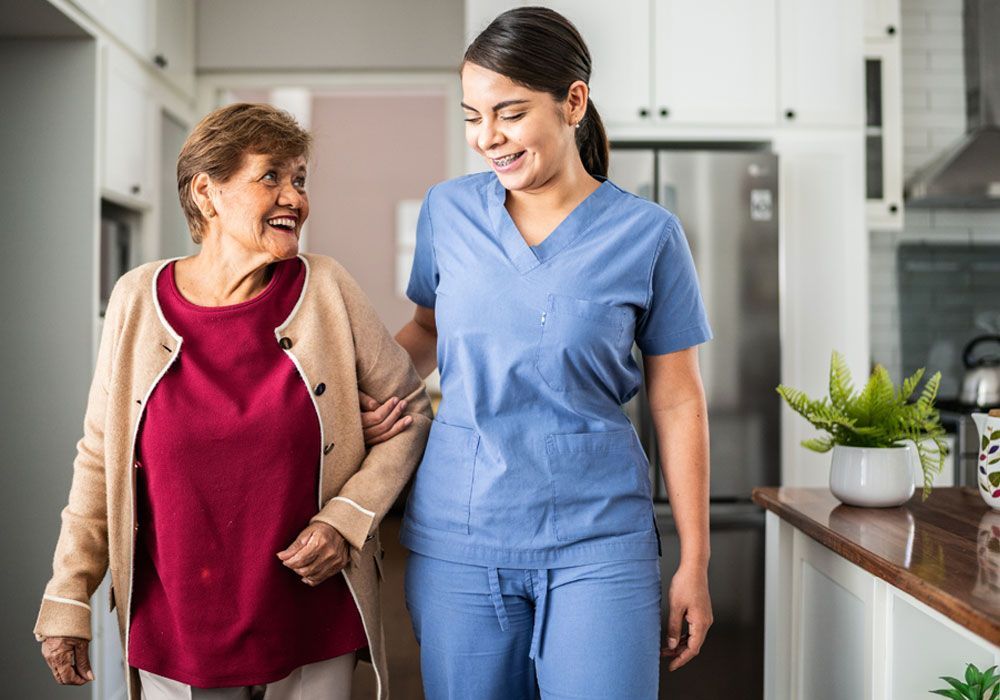 Two women walking together in a bright kitchen, smiling and talking, one in scrubs and one in a red top and beige cardigan