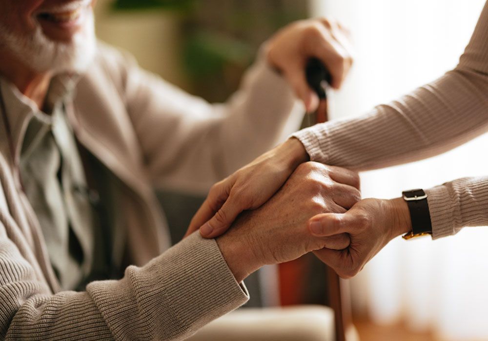 Two hands clasped in support, with an elderly person holding a cane in the background