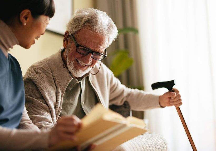 Two people reading together on a couch, one holding a book and the other smiling with a cane.