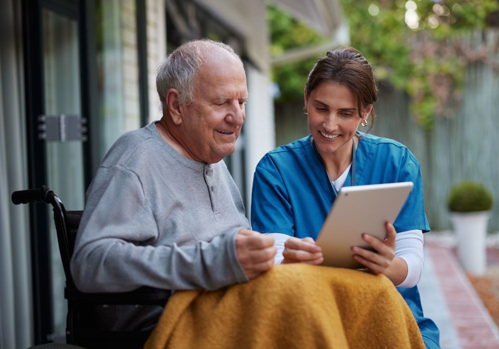 Caregiver showing a tablet to an older adult on a porch, both smiling and looking at the screen