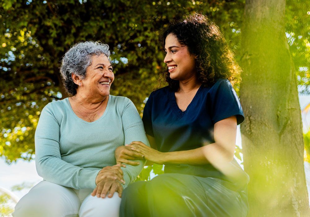 Caregiver showing a tablet to an older man seated outdoors, both smiling under a blanket