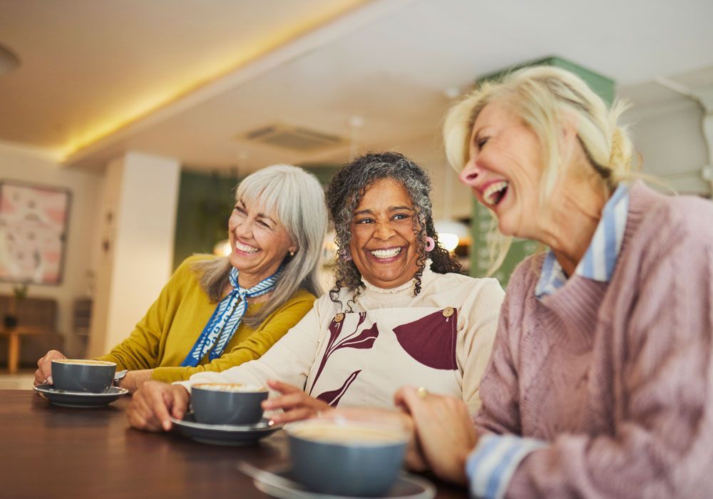 Two smiling women sitting close together indoors, one in pink and one in light blue, sharing a laugh.