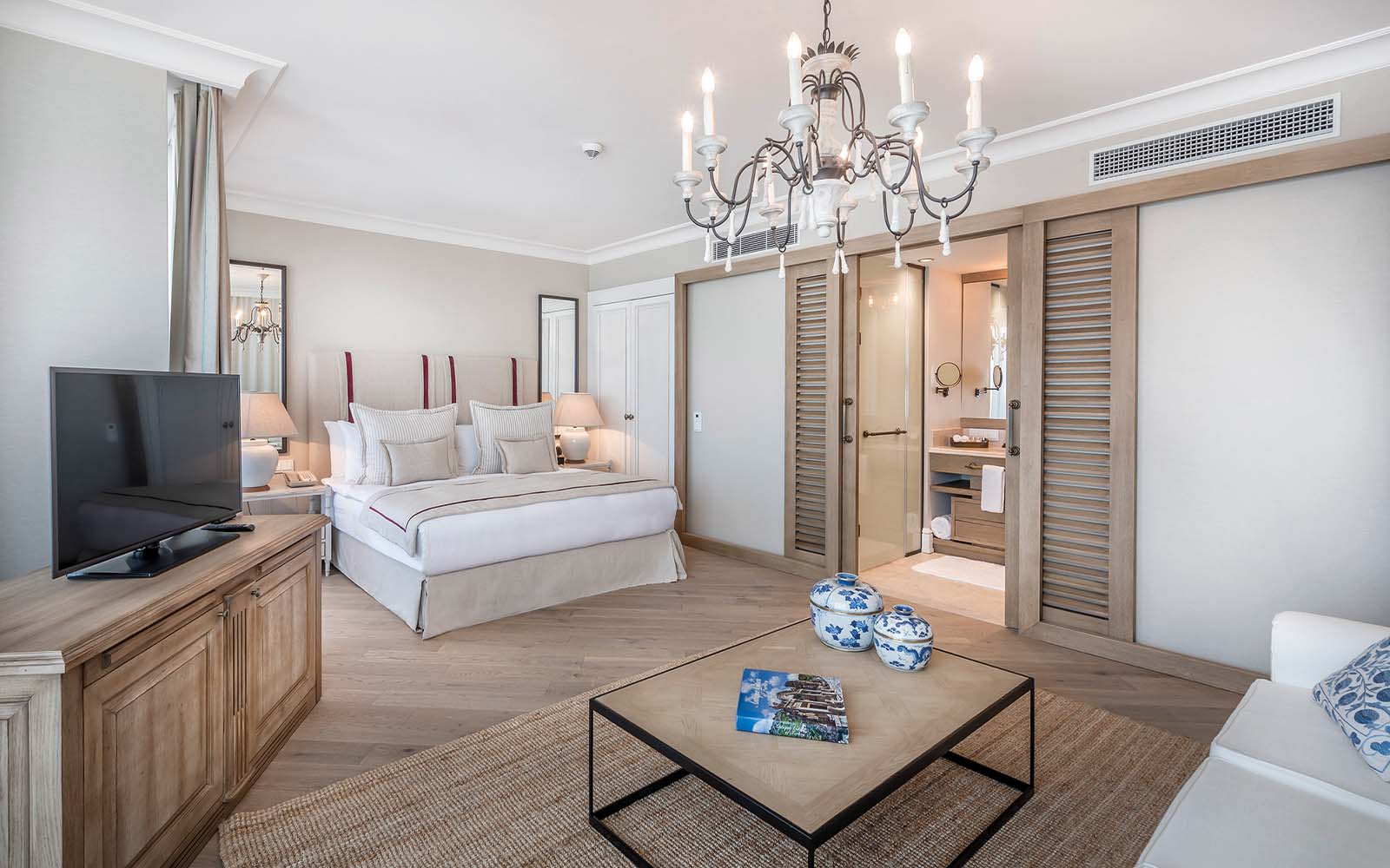 A hotel room with a bed , television , coffee table and chandelier.