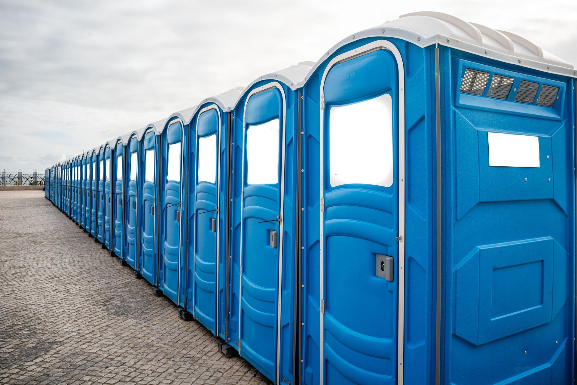 Long row of blue portable toilets with white roofs on a paved outdoor area.