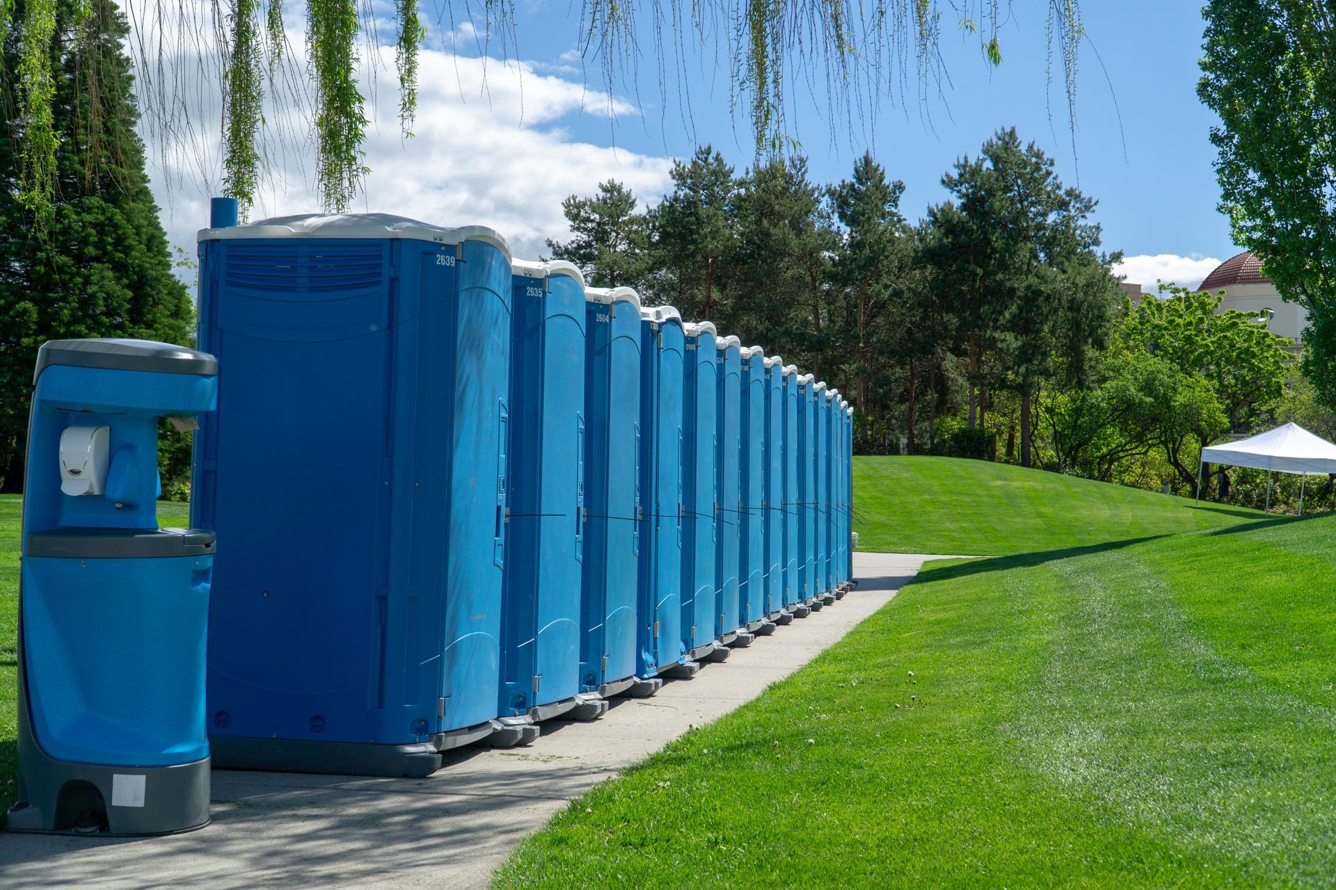 Blue portable toilets lined along a walkway in a landscaped park setting.