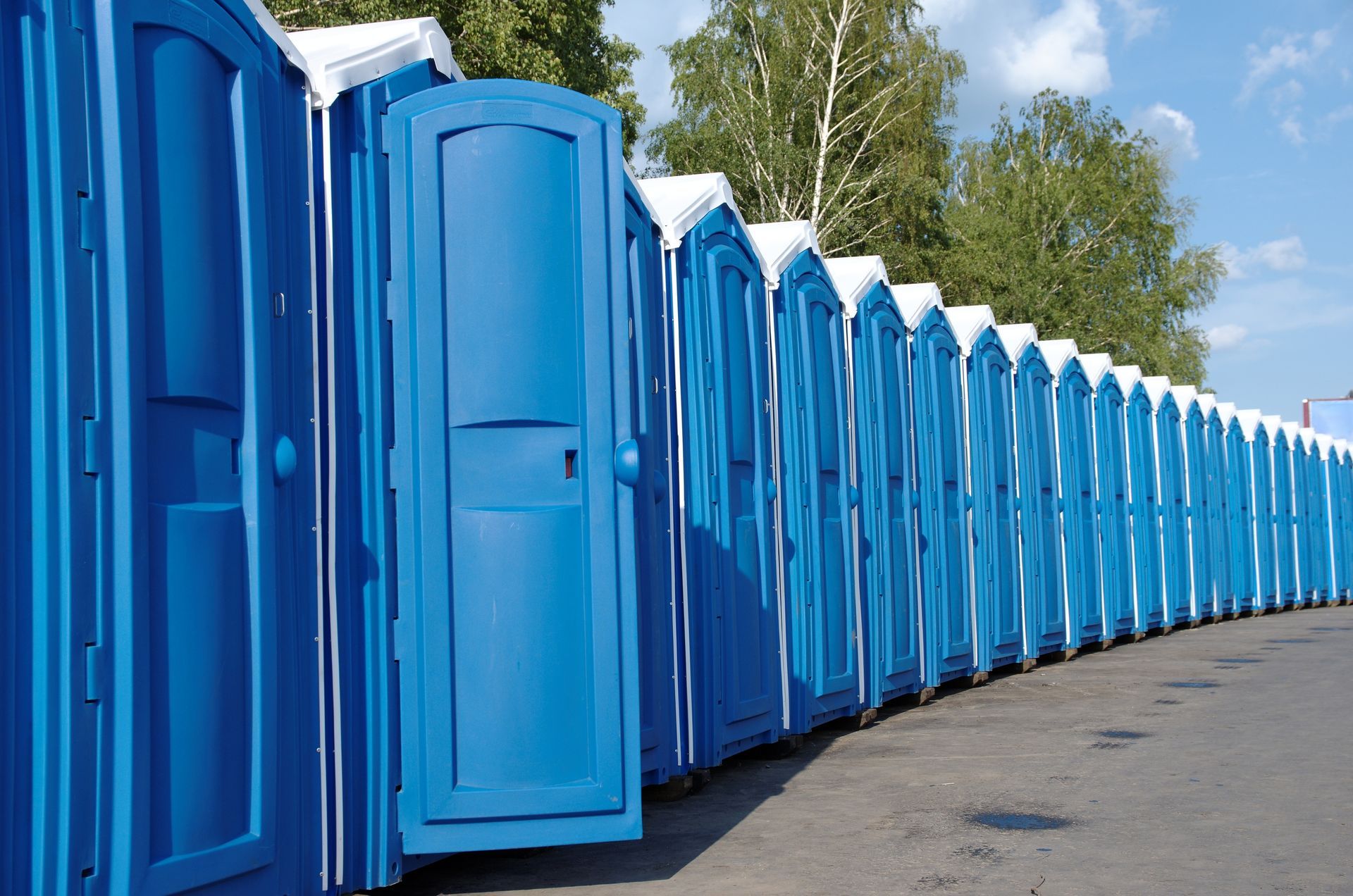 Row of blue portable toilets lined up outdoors on a paved surface. Row of blue portable toilets lined up outdoors on a paved surface.