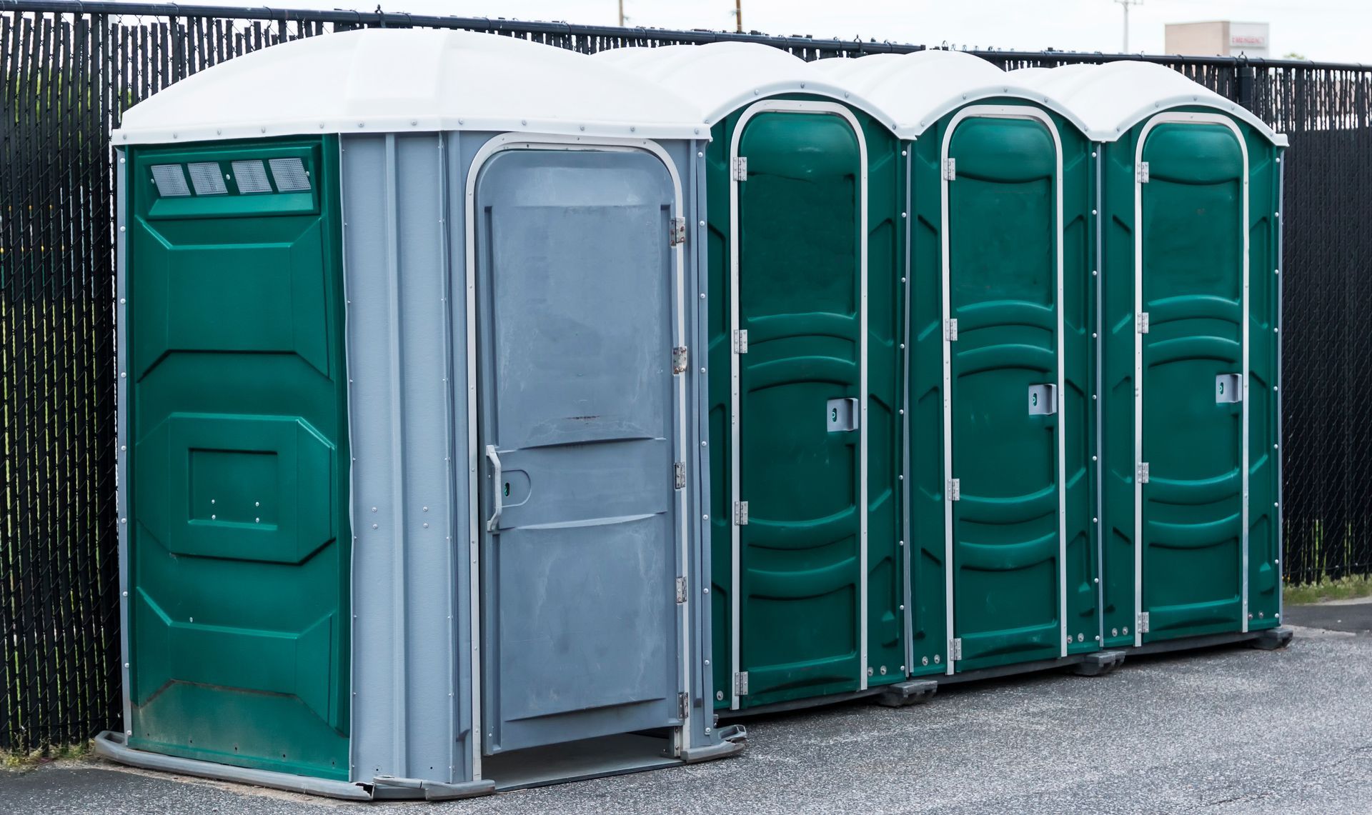 Photograph of four portable toilets lined up outdoors on a paved surface with a black fence in the background.