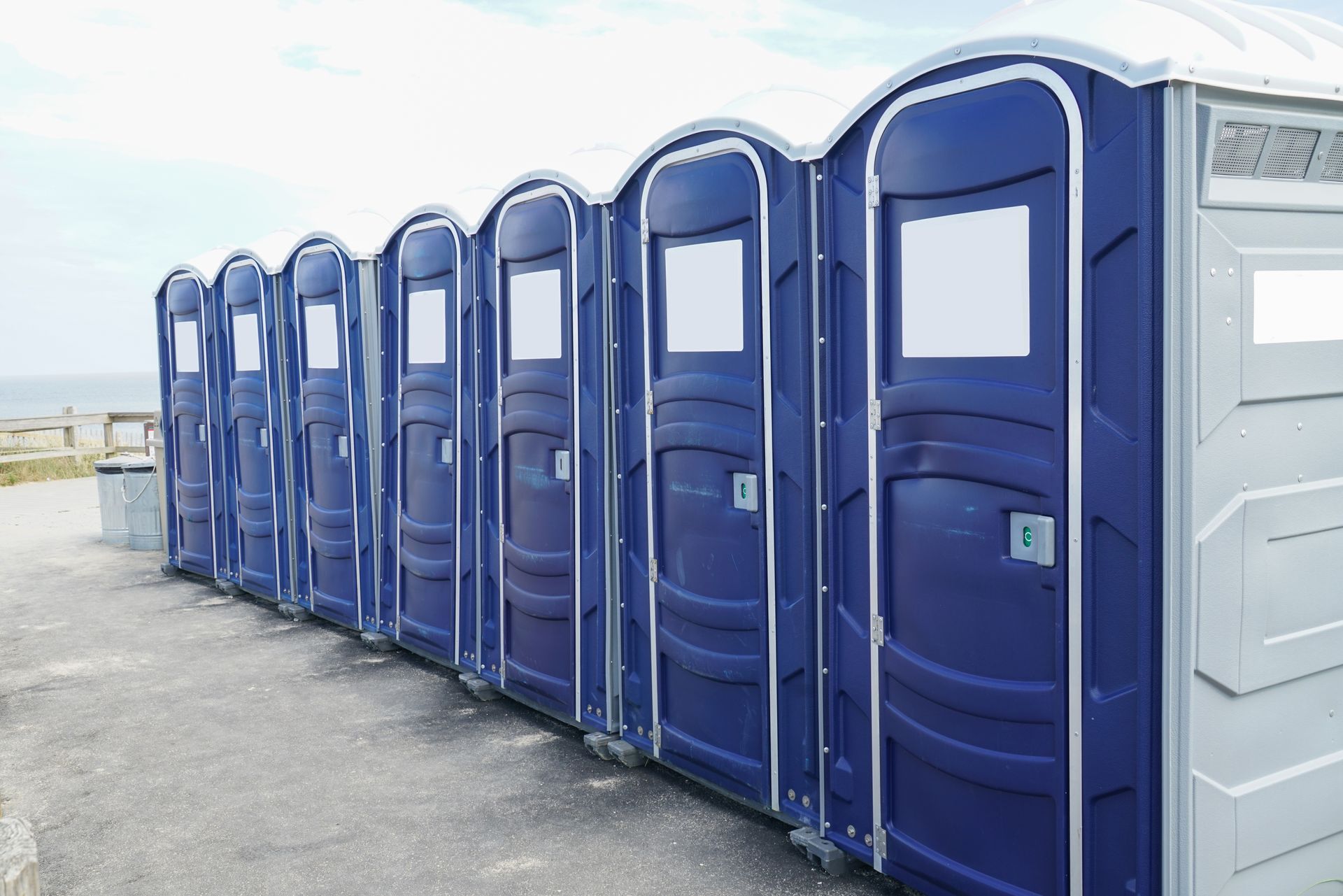A row of blue portable toilets lined up outdoors on a paved surface near a wooden fence.