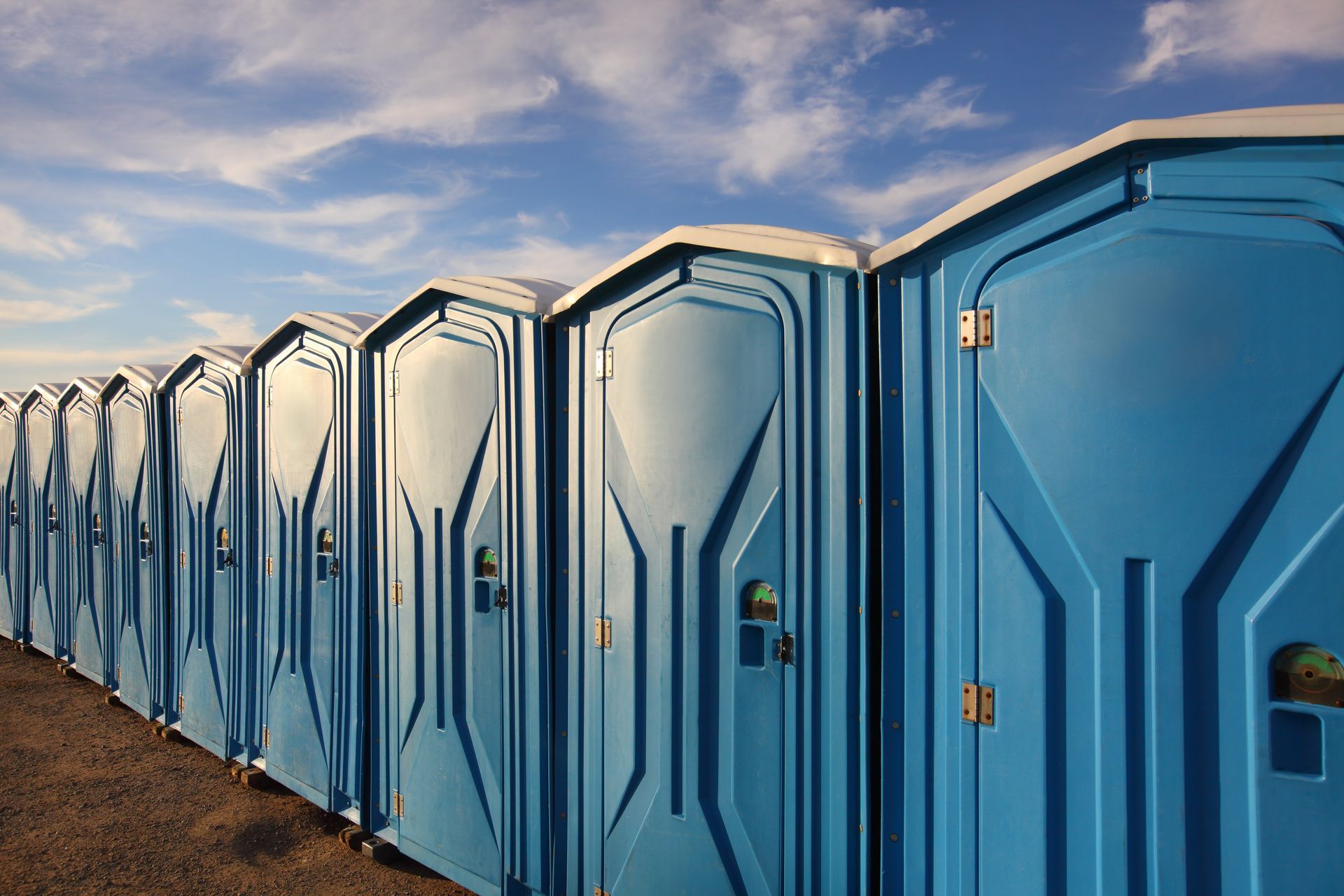 Blue portable toilets lined up outdoors beneath a partly cloudy sky.