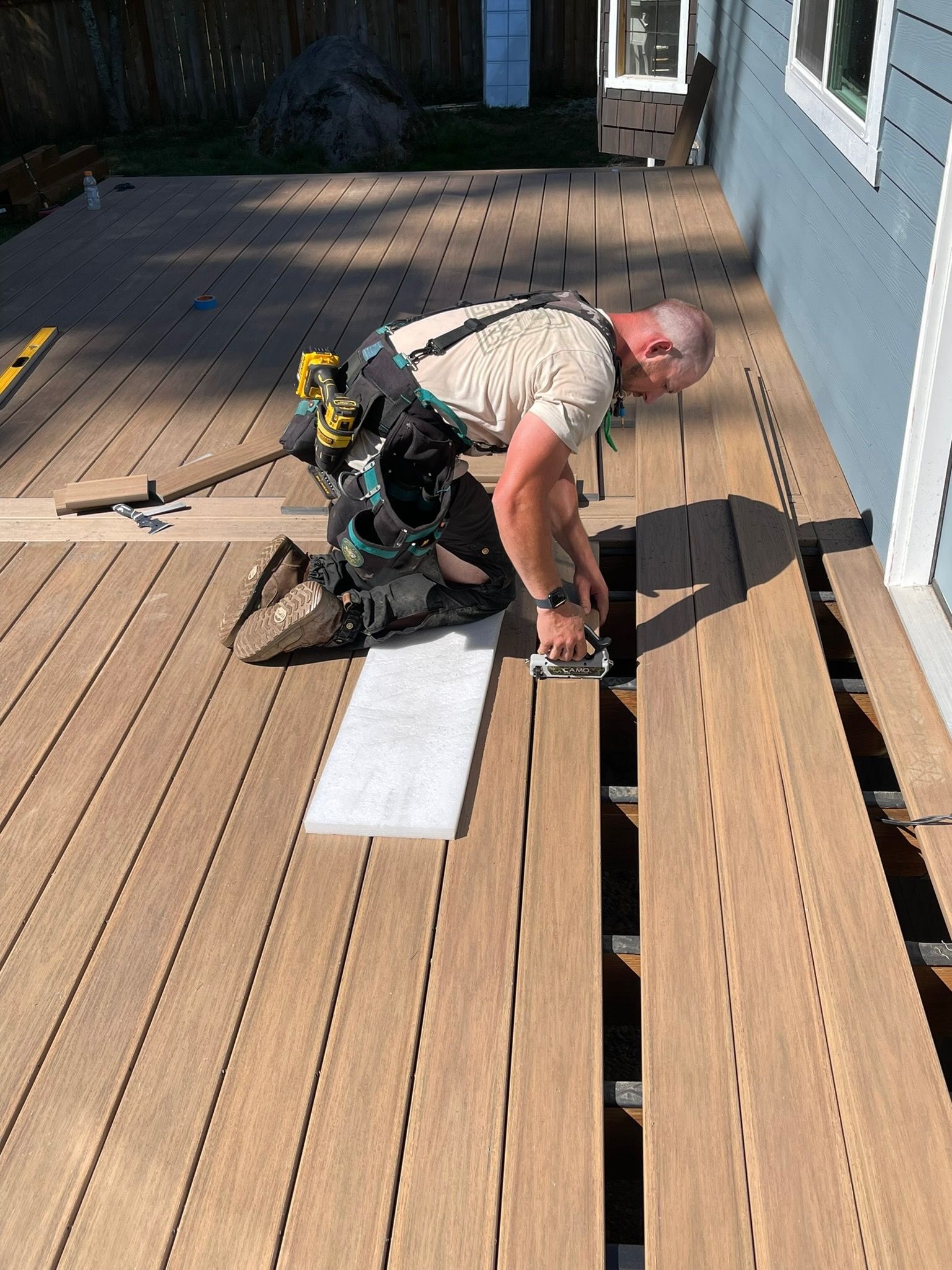 A construction worker kneeling on a new composite deck, installing planks next to a light-blue house.