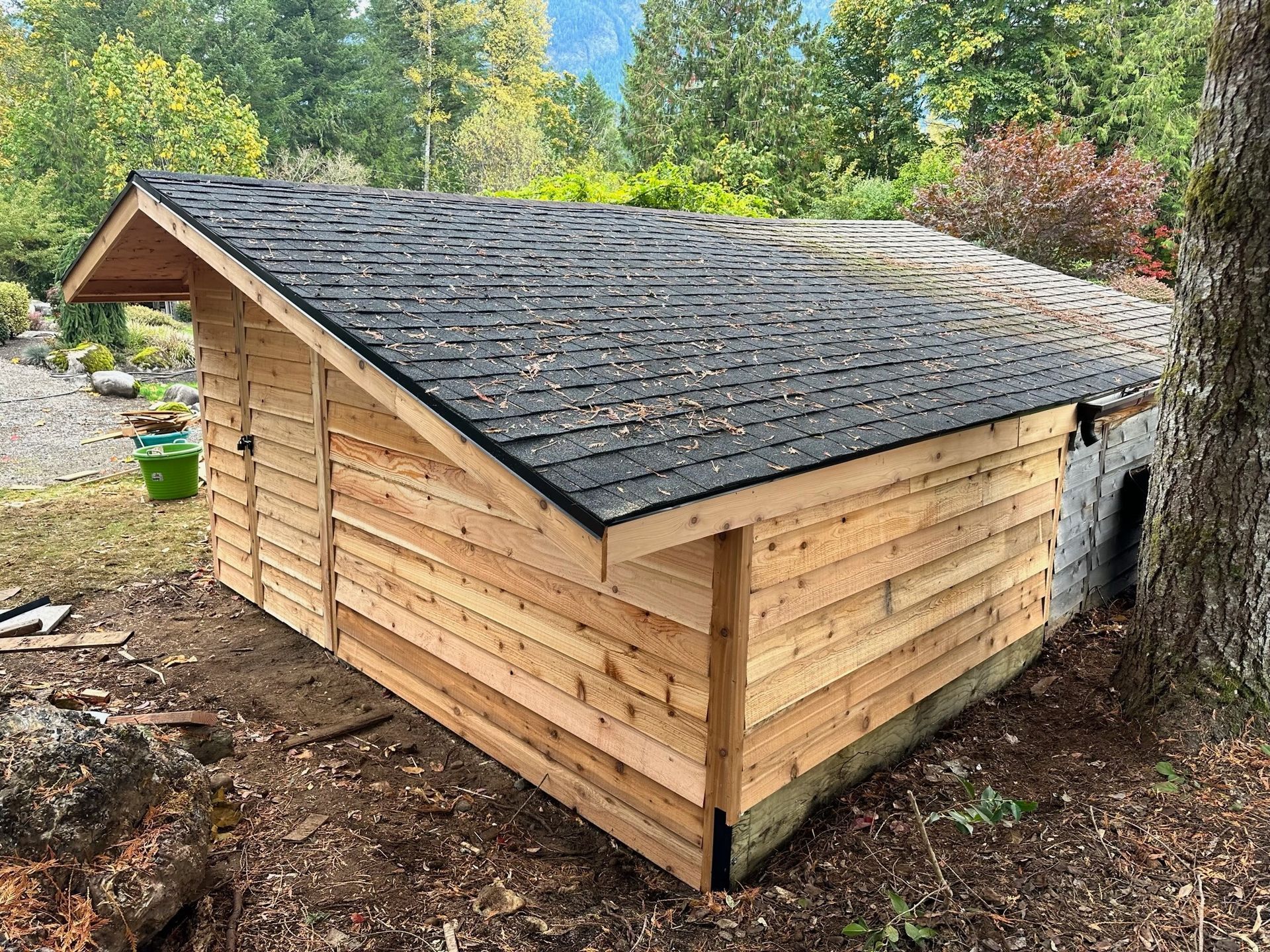 A wooden shed with a shingled roof, built against a stone structure in a wooded, outdoor setting.