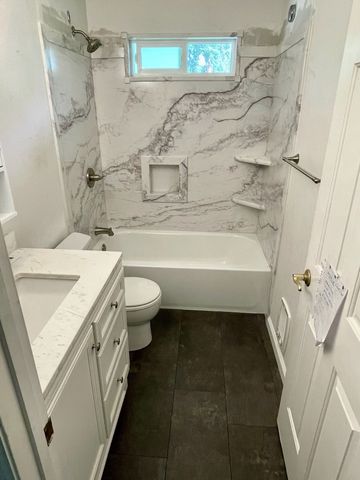 A bright bathroom featuring a white vanity, a toilet, and a bathtub with marble-patterned tile walls and dark tile floors.