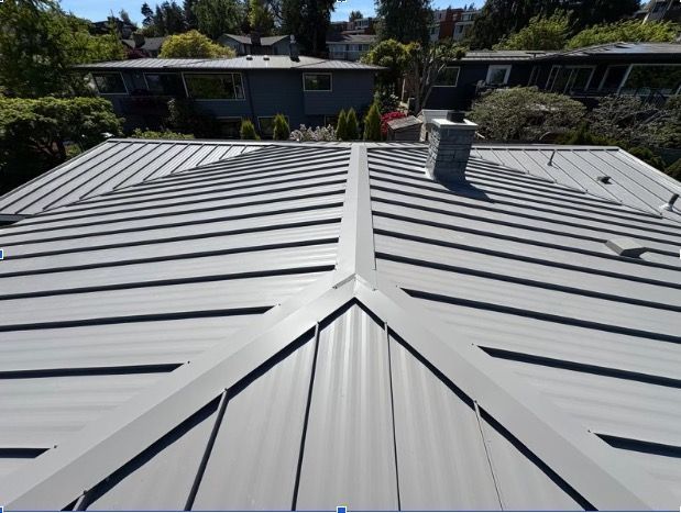 A worker installs dark metal roofing panels over a blue underlayment on a house roof.