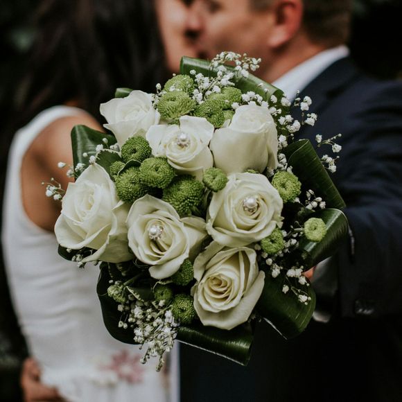 A bride and groom are kissing while the bride is holding a bouquet of white roses and greenery.