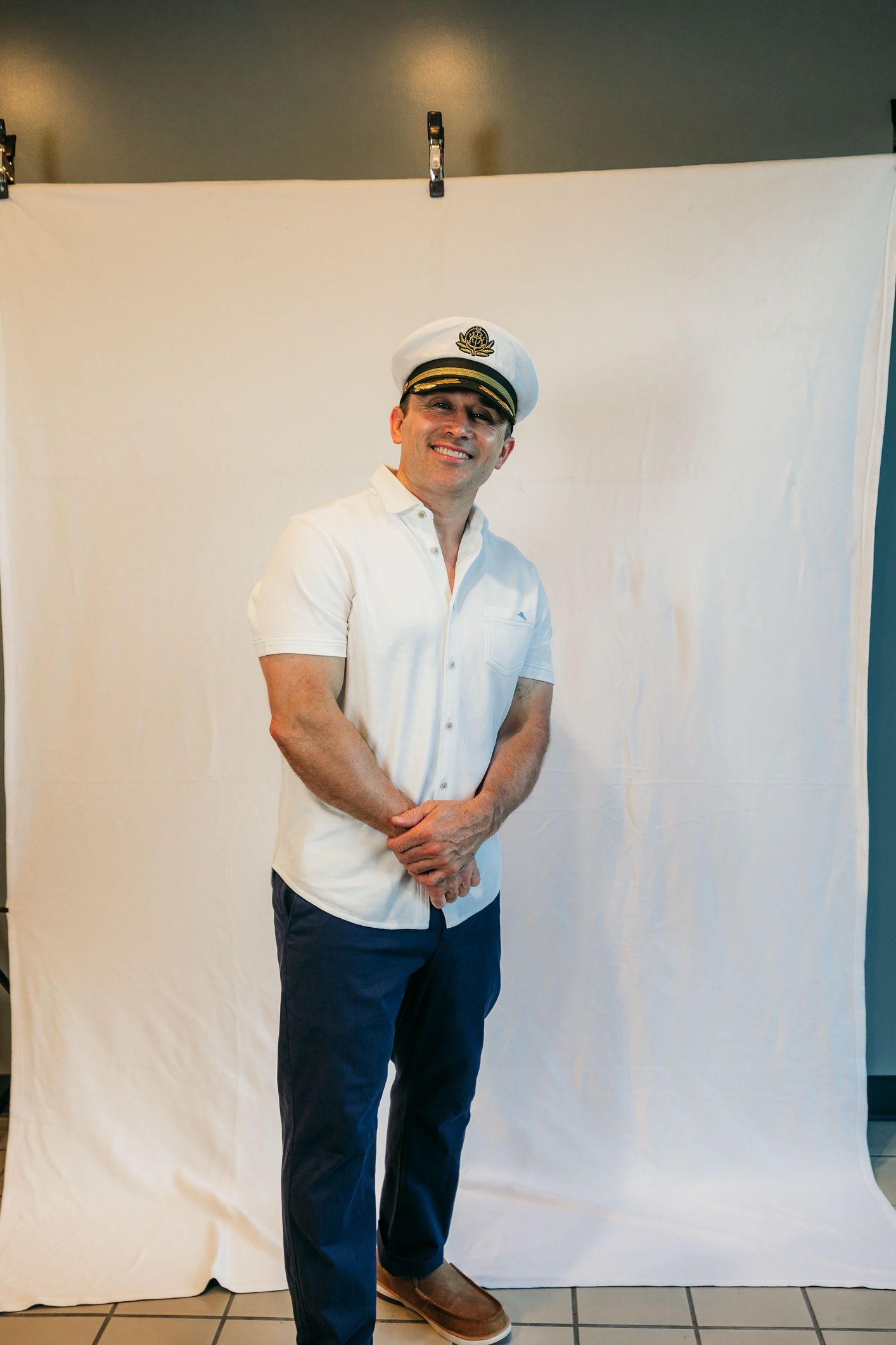 Man in white shirt and blue pants posing in front of a white backdrop, wearing a captain’s hat.