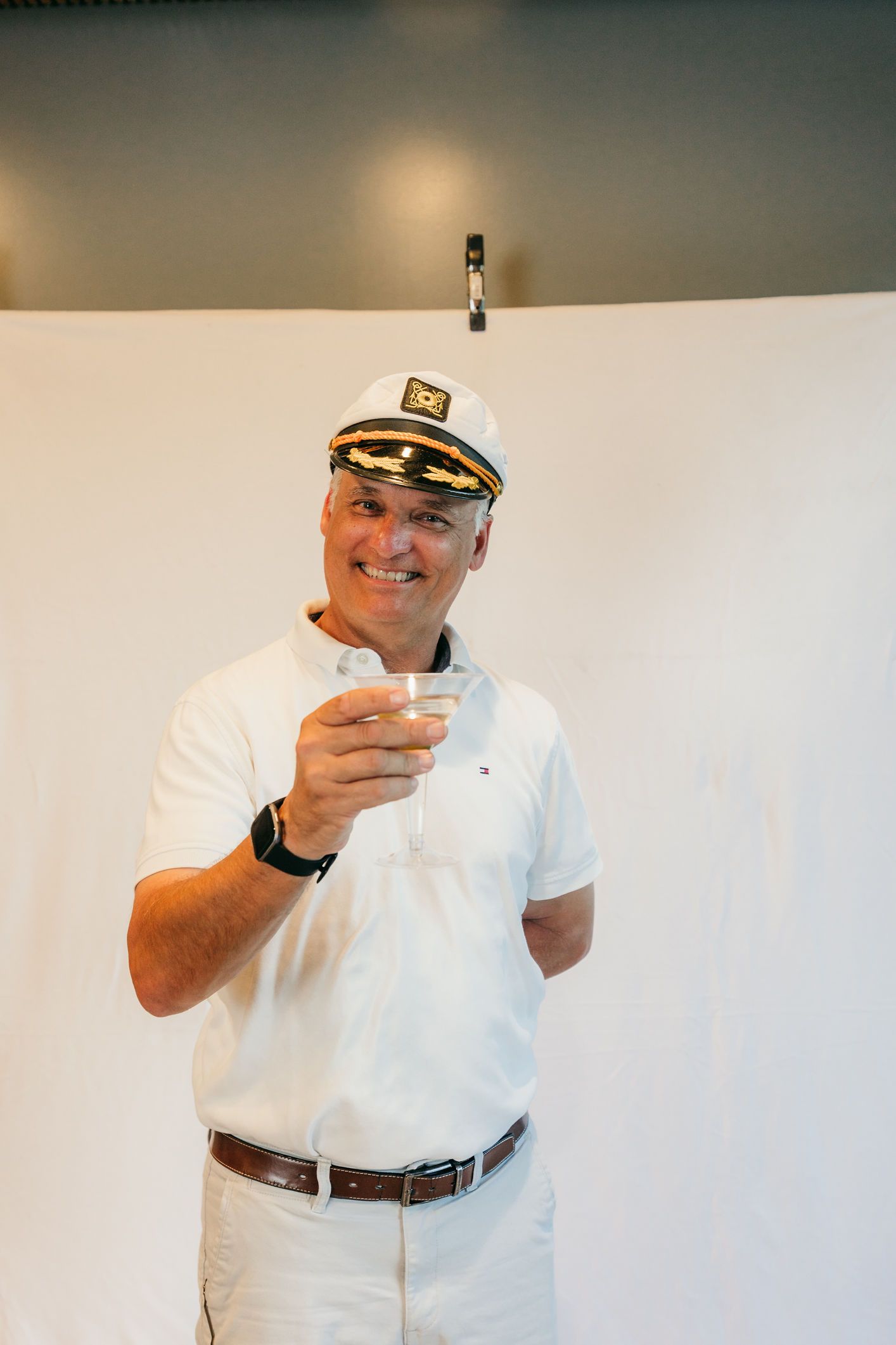 Man in white shirt and captain’s hat holding up a drink, standing in front of a plain wall.