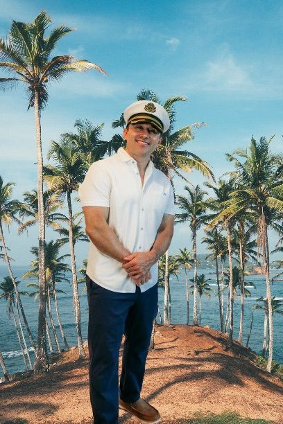 Man in white shirt and captain’s hat posing on a tropical hill with palm trees and ocean behind him