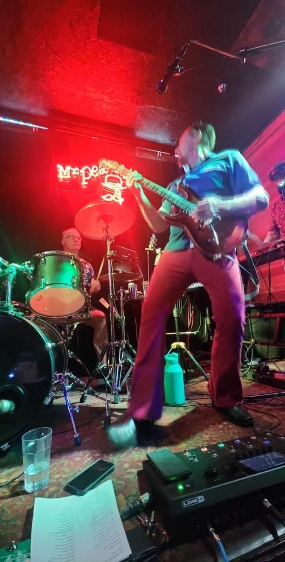 Musician singing and playing guitar on a red-lit stage with drums behind him